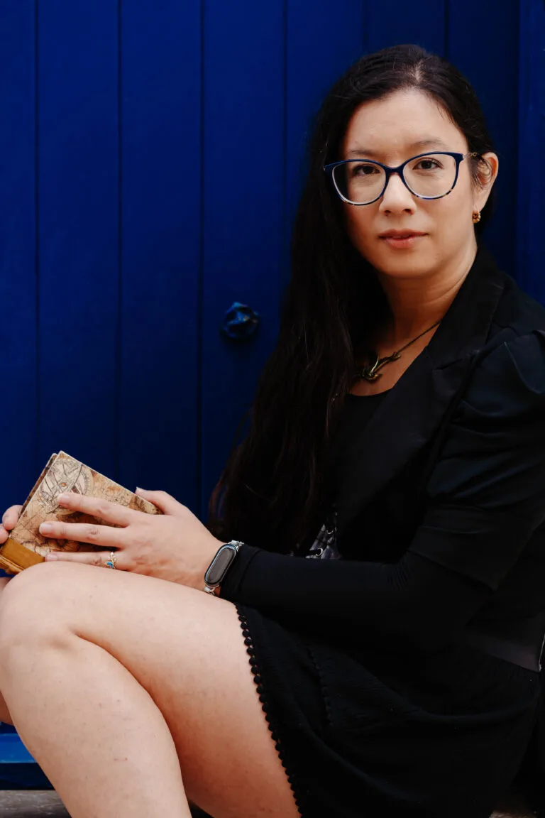 Photo of Amy V Borg. She is a dark haired woman with glasses, sitting in a doorway against a dark blue door with a notebook in hand.